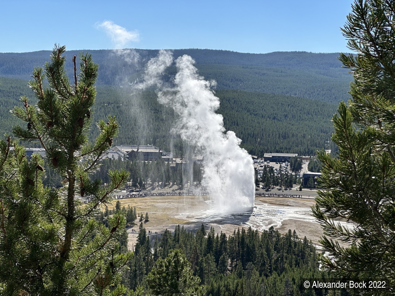 View from above of old faithful erupting, surrounded by a crowd